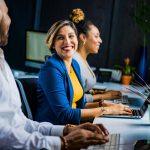 Three diverse professionals working and smiling at office desks, fostering teamwork and collaboration.
